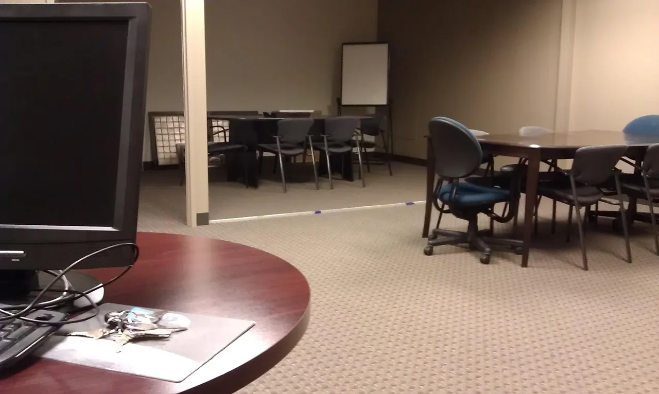 Dated office meeting room with mismatched chairs, an old boxy monitor and a whiteboard on a stand