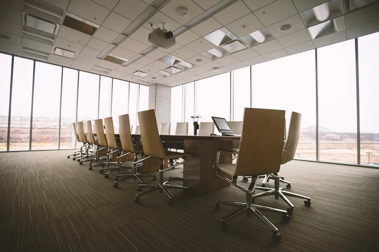Empty glass-walled boardroom with a long wooden table and a row of vacant office chairs beside floor-to-ceiling windows