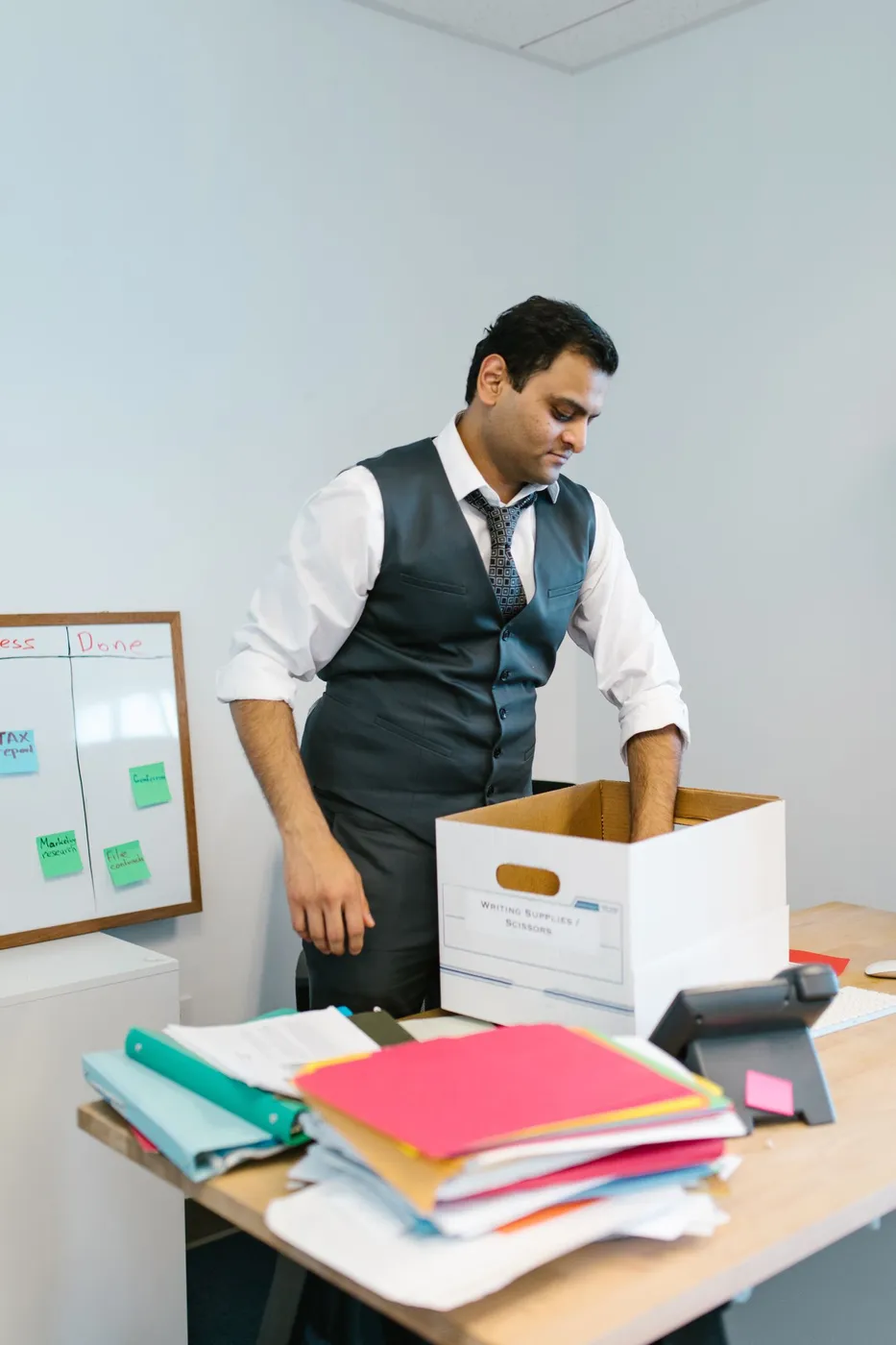 Man in a grey waistcoat packing an archive box on a desk stacked with coloured folders and a desk phone