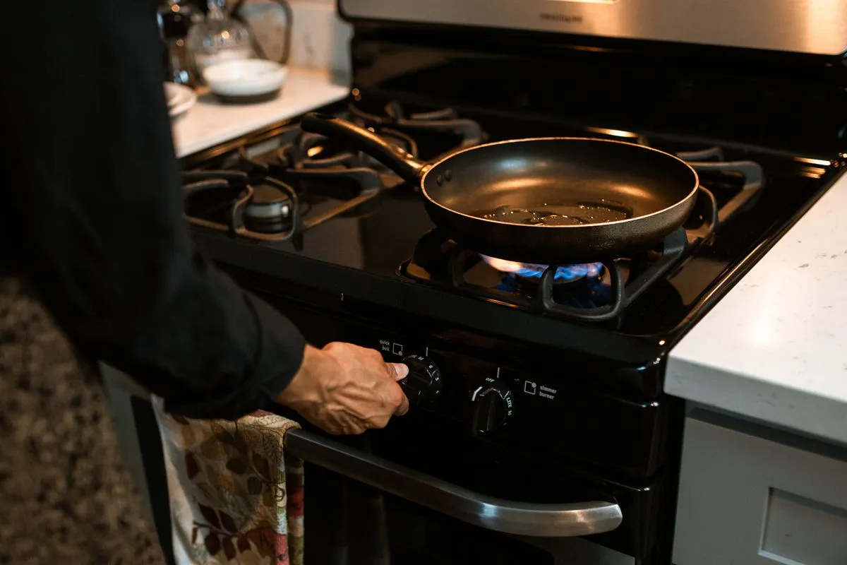 Hand turning a control knob beside a lit gas burner with a frying pan on a black stovetop