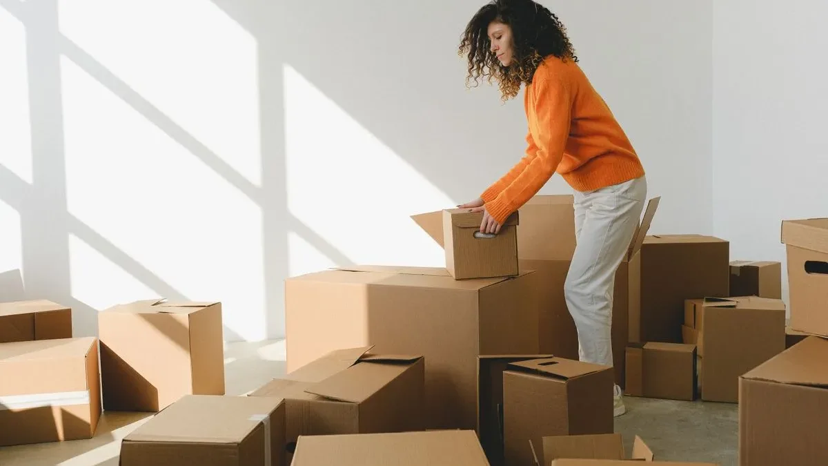 Woman in an orange jumper leaning over one of many cardboard moving boxes spread across a sunlit, otherwise empty room