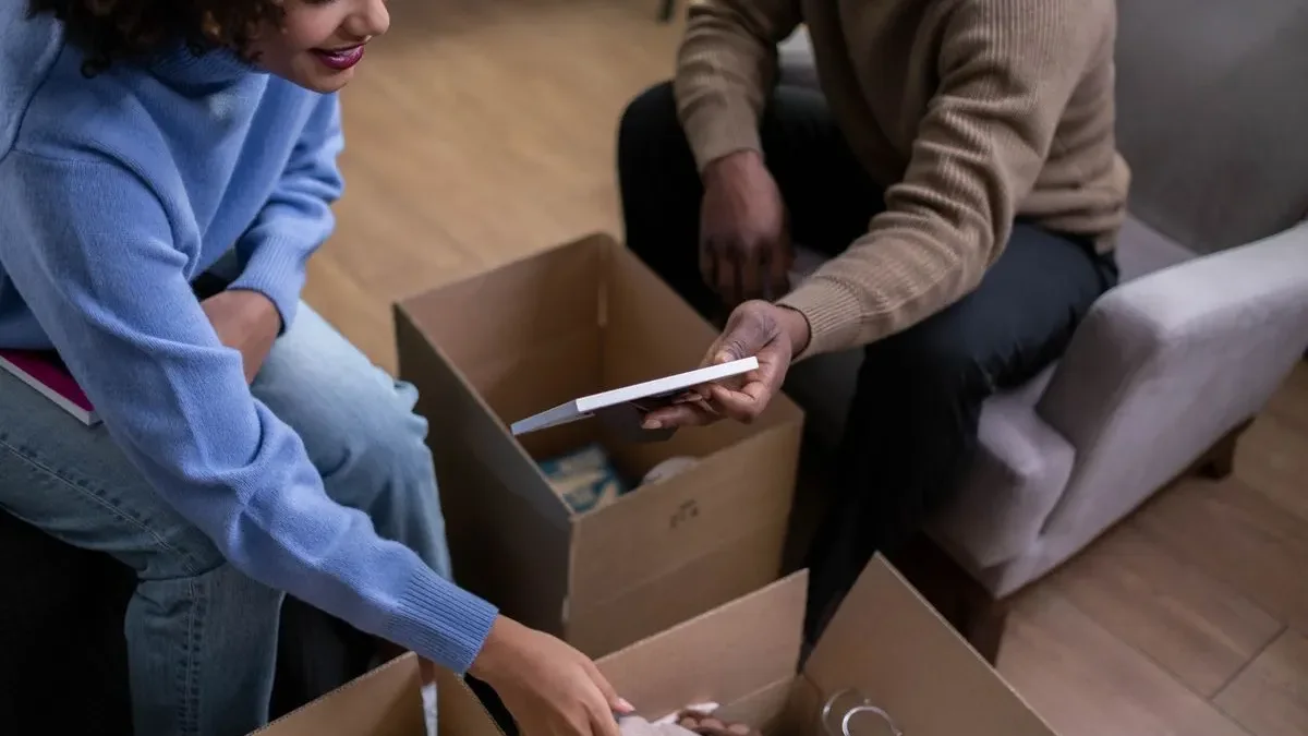 Woman in a blue jumper and man in a brown jumper sorting through open cardboard boxes, one holding up a flat hardback item while deciding whether it stays