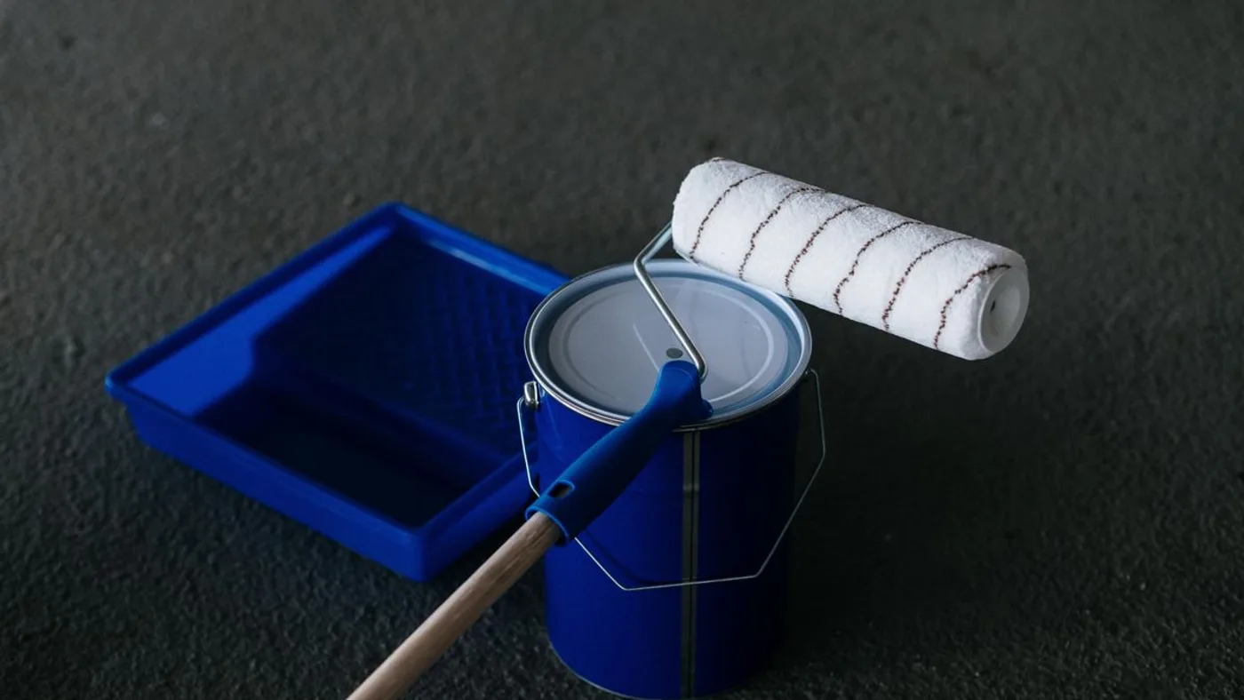 Unopened blue paint tin with a roller resting across the lid and an empty paint tray behind it on a dark floor