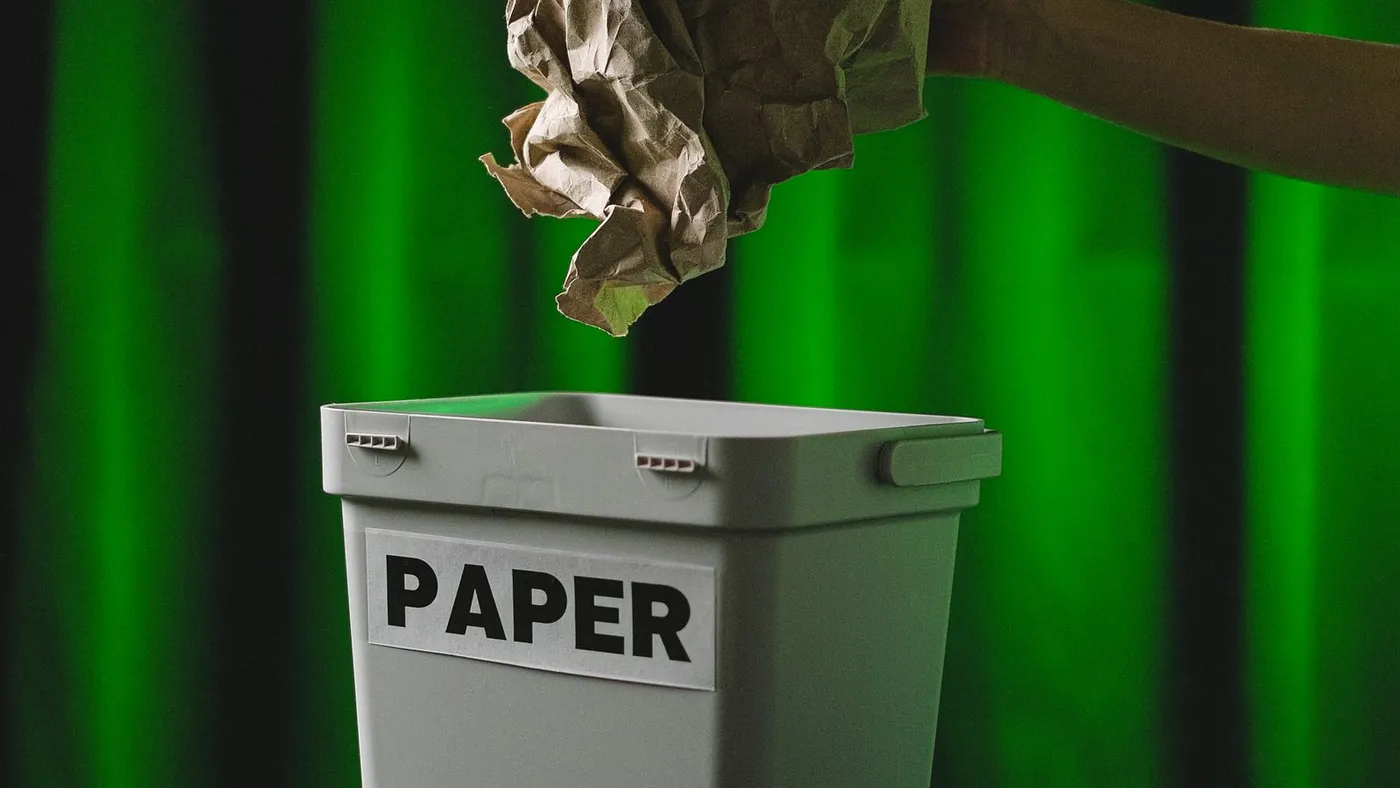Hand dropping crumpled brown paper into a grey bin labelled PAPER against a green lit backdrop