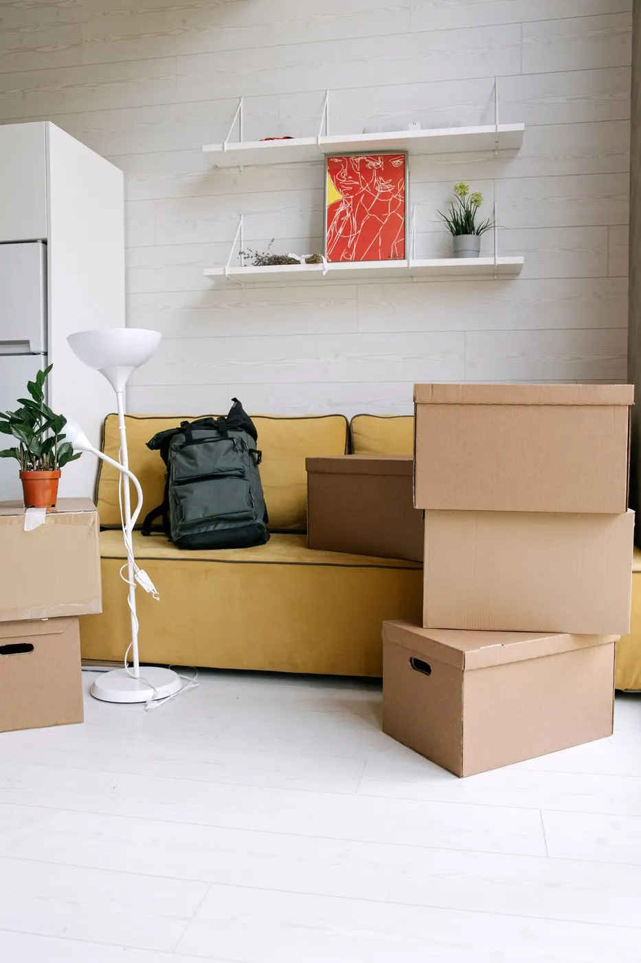Living room with a yellow sofa, a backpack and stacked cardboard boxes while wall shelves and art remain in place