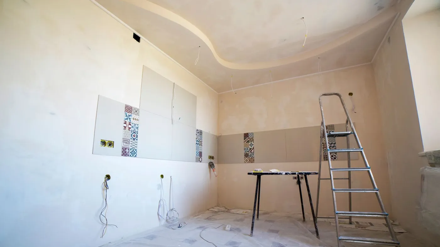Kitchen mid-renovation with patterned tile squares half-fitted on the wall, a stepladder and a folding table covered in dust sheets