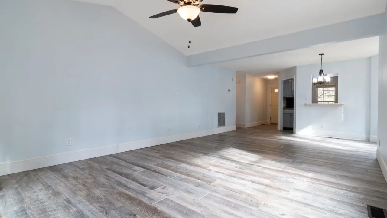 Empty living room with pale walls, a ceiling fan and grey wood-effect plank flooring, opening onto a hallway and a dining area