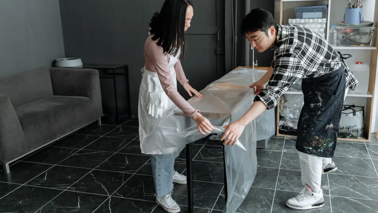 Two people in aprons wrapping a table in clear plastic sheeting on a dark marble-effect tiled floor next to a grey sofa