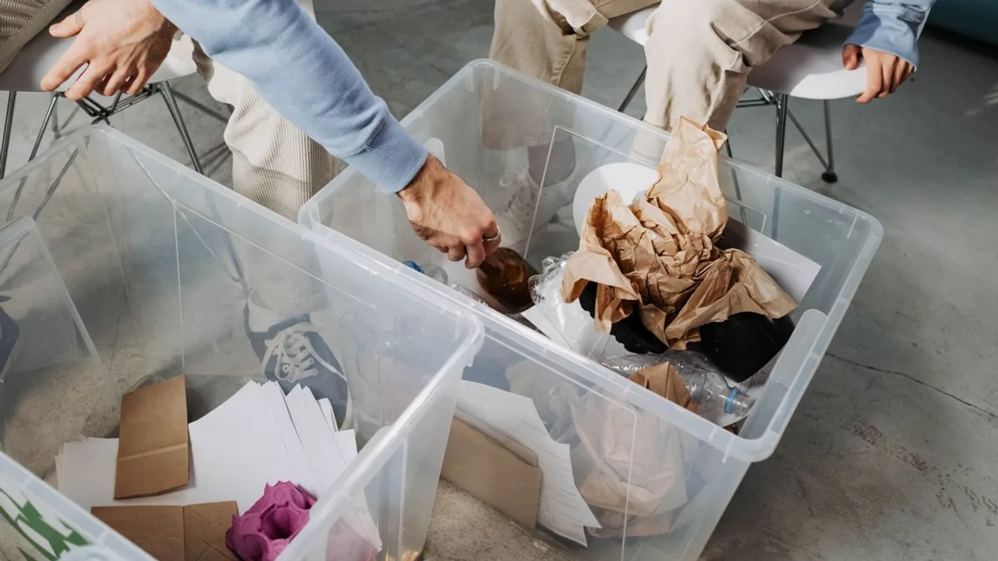Two people crouching over large clear plastic crates, sorting paper, bottles and packaging into separate containers