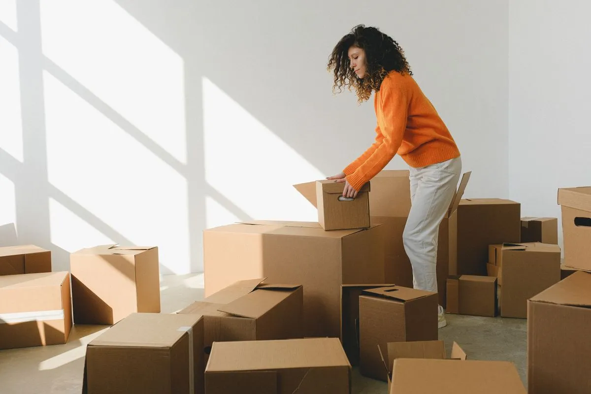 Woman in an orange sweater packing a cardboard box in a sunlit, mostly empty room during a house move