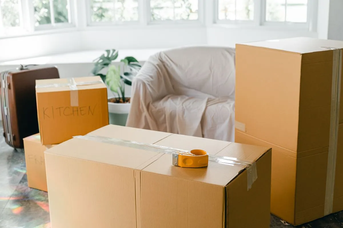 Cardboard boxes labelled Kitchen stacked beside a dust-sheet-covered armchair in a Dubai apartment being packed for a move