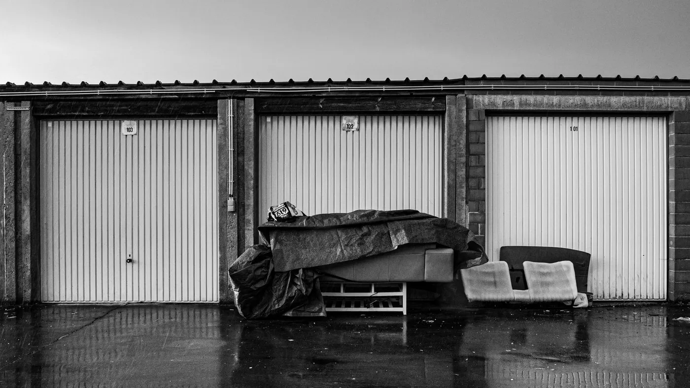 Black and white photo of a sofa wrapped in a tarpaulin cover standing outside a row of numbered storage garage doors