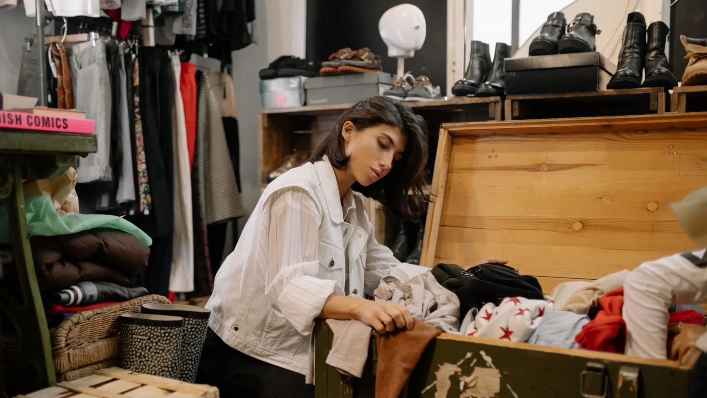 Woman kneeling beside an open wooden storage trunk filled with folded clothing, surrounded by shoes, boots and a clothes rack
