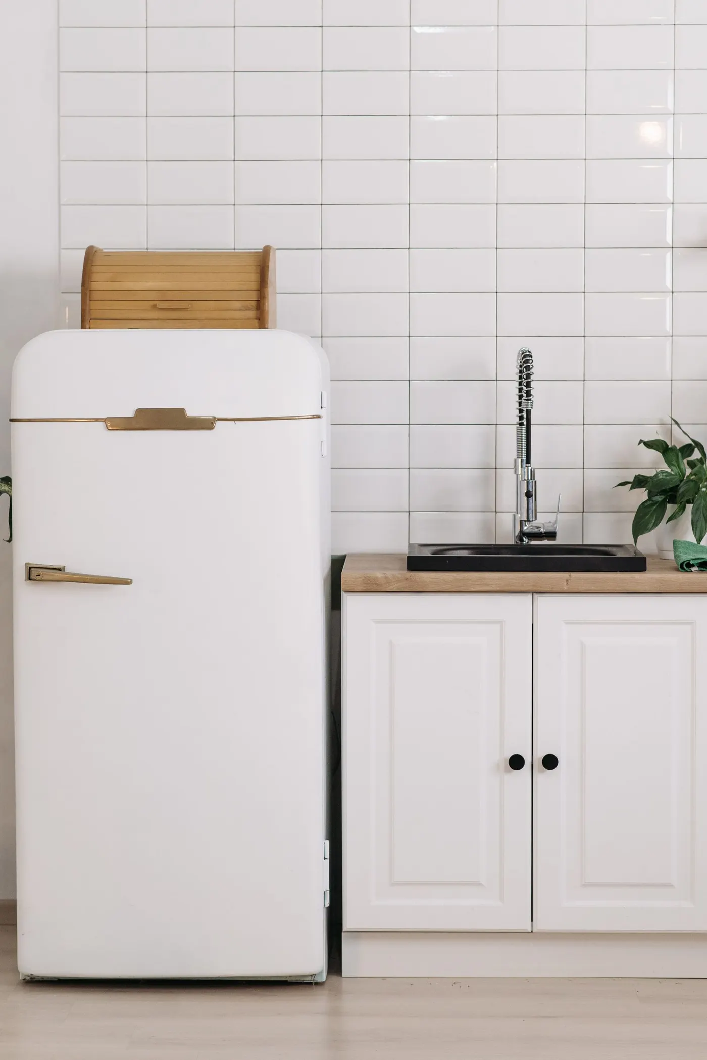 White retro-styled fridge with a brass handle standing beside a white kitchen cabinet with a wooden worktop, black sink and white tiled wall