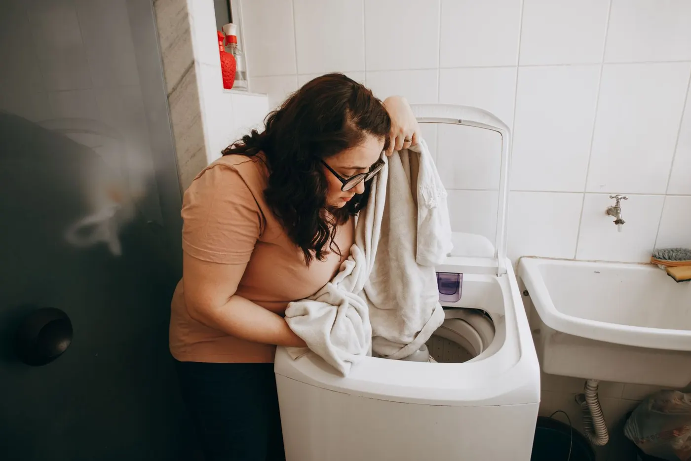 Woman in an orange t-shirt and glasses loading pale towels into an open top-loading washing machine in a white tiled utility area beside a sink