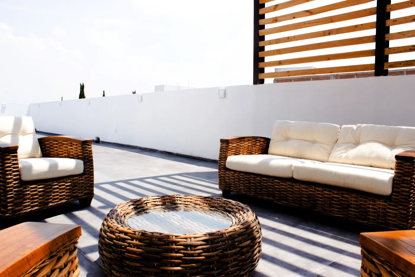 Sunlit rooftop terrace with brown wicker sofas and cream cushions beside a low white parapet wall and a timber slatted screen