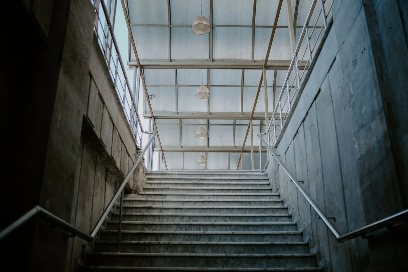 Wide bare concrete staircase between metal handrails leading up toward a glazed roof inside a building