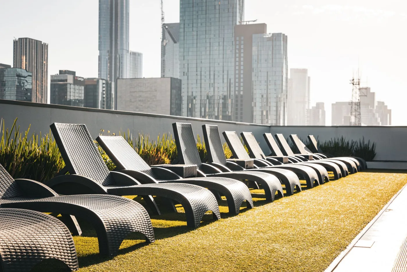 Row of dark rattan sun loungers on a high roof terrace with a low perimeter wall and city towers rising behind