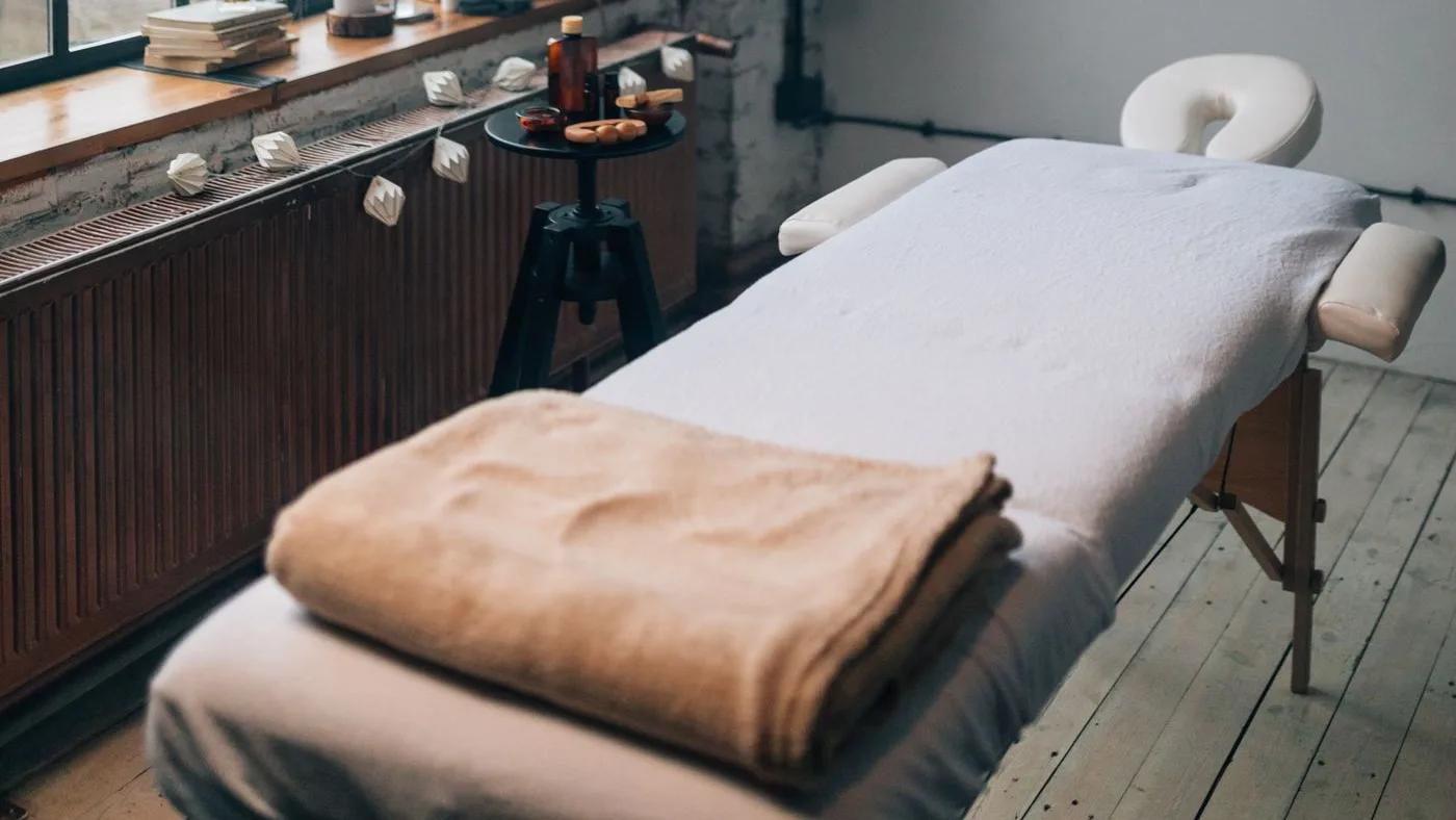Spa treatment room with a padded massage couch dressed in white towelling, a folded tan blanket and a small round side table by a tall window
