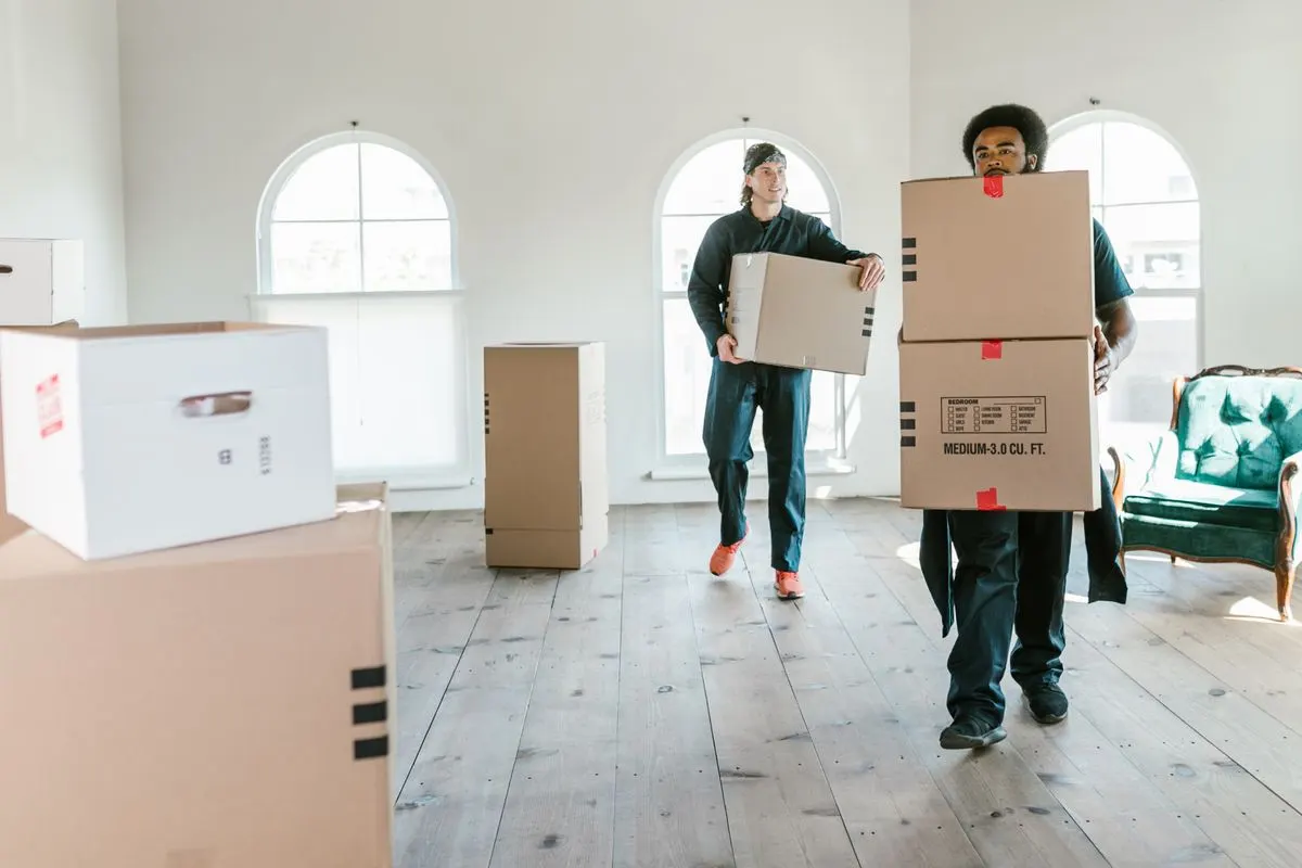 Two movers in overalls carrying labelled cardboard boxes through a bright, mostly empty room with arched windows