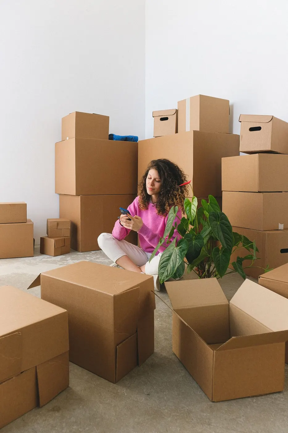 Woman in a pink sweater sitting on the floor among stacked cardboard boxes while using her phone