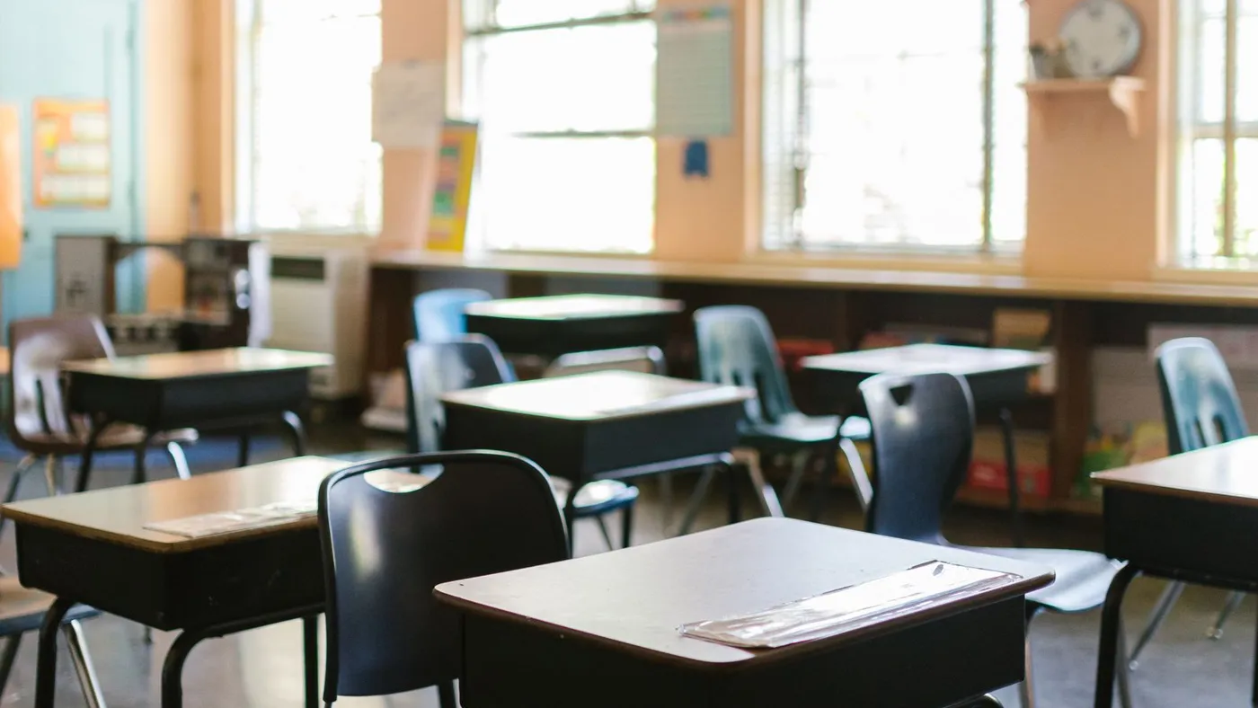 Sunlit classroom with rows of individual wooden lift-lid desks paired with black and blue moulded plastic chairs