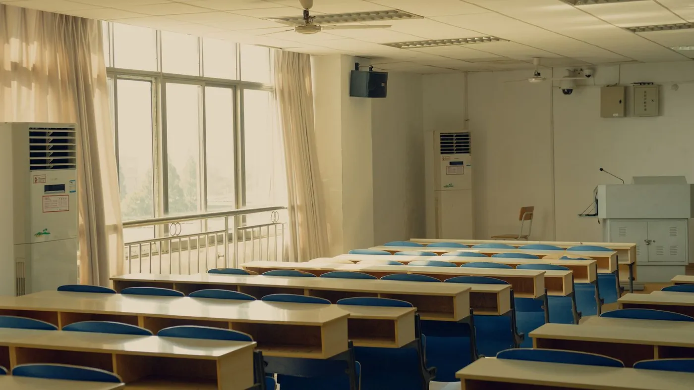 Empty lecture room with rows of pale wooden bench desks and blue folding seats beside tall curtained windows and a lectern