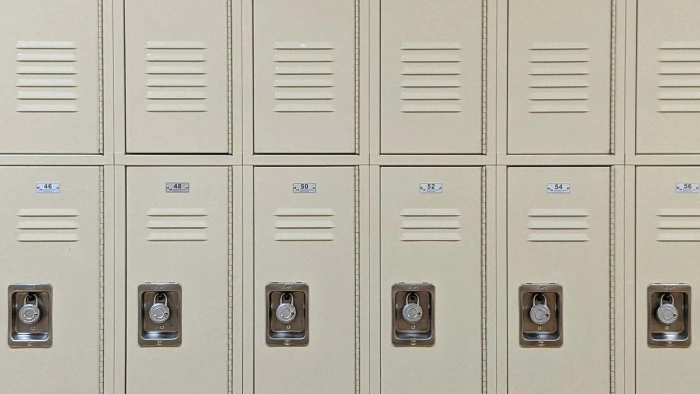 Row of pale beige metal school lockers with combination locks and louvred vents above a wooden floor and dark skirting