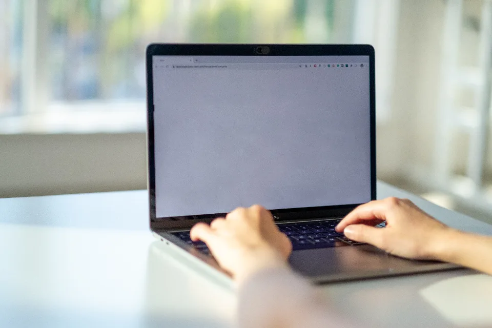 Person typing on a laptop keyboard at a home desk with a blank browser window open