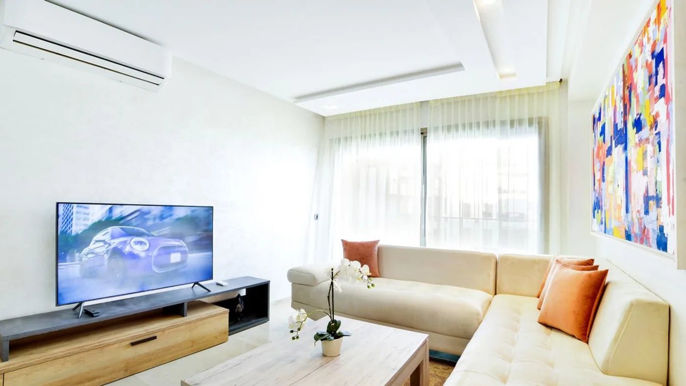 Furnished apartment living room with a wall-mounted split air conditioning unit, a large television on a low media unit and a cream corner sofa