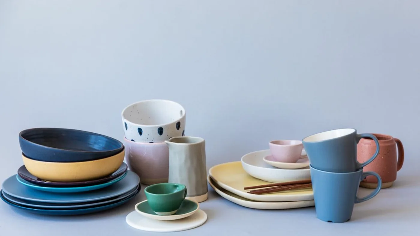 Mixed set of household crockery grouped together: stacked plates, bowls and mugs in blue, pink, cream and grey against a plain background