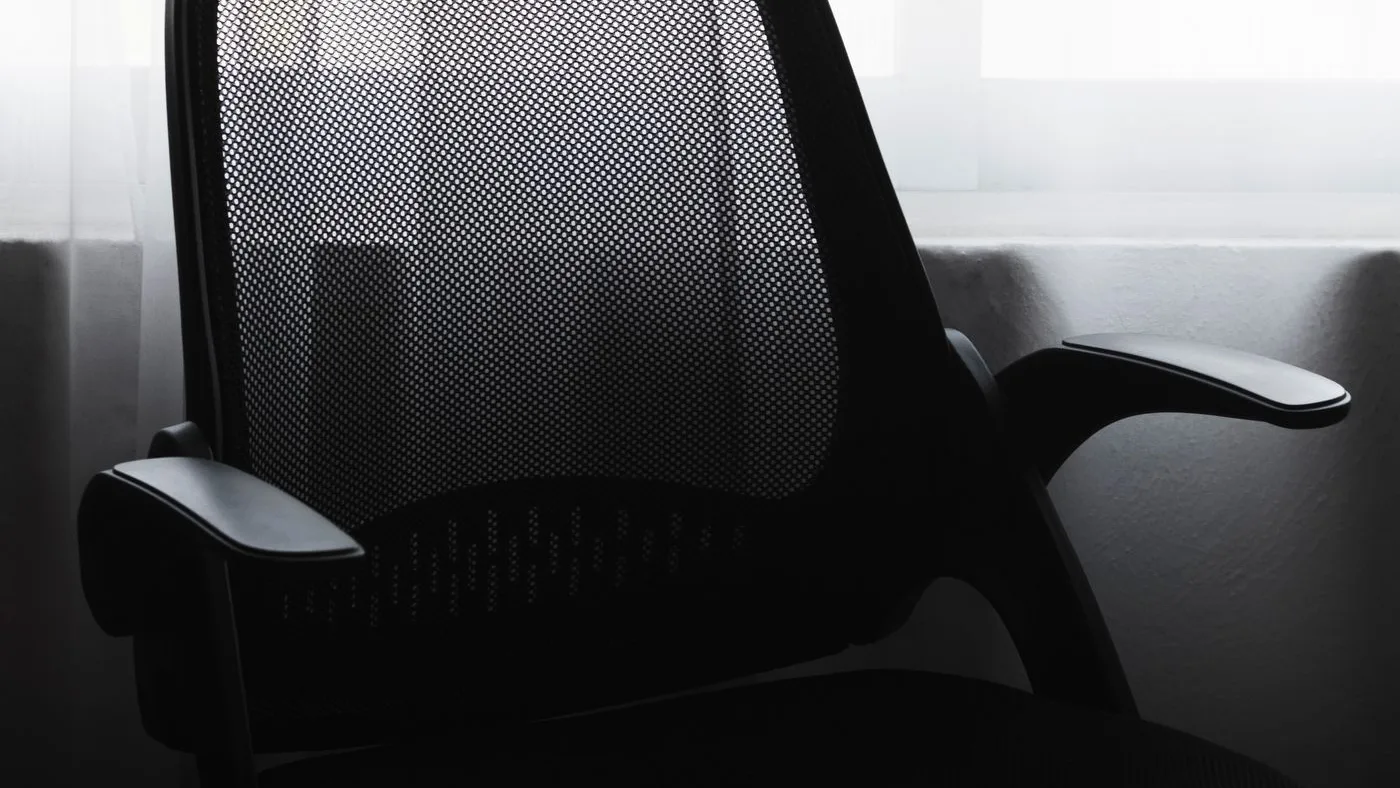 Single black mesh-backed task chair with padded armrests silhouetted against a bright curtained window