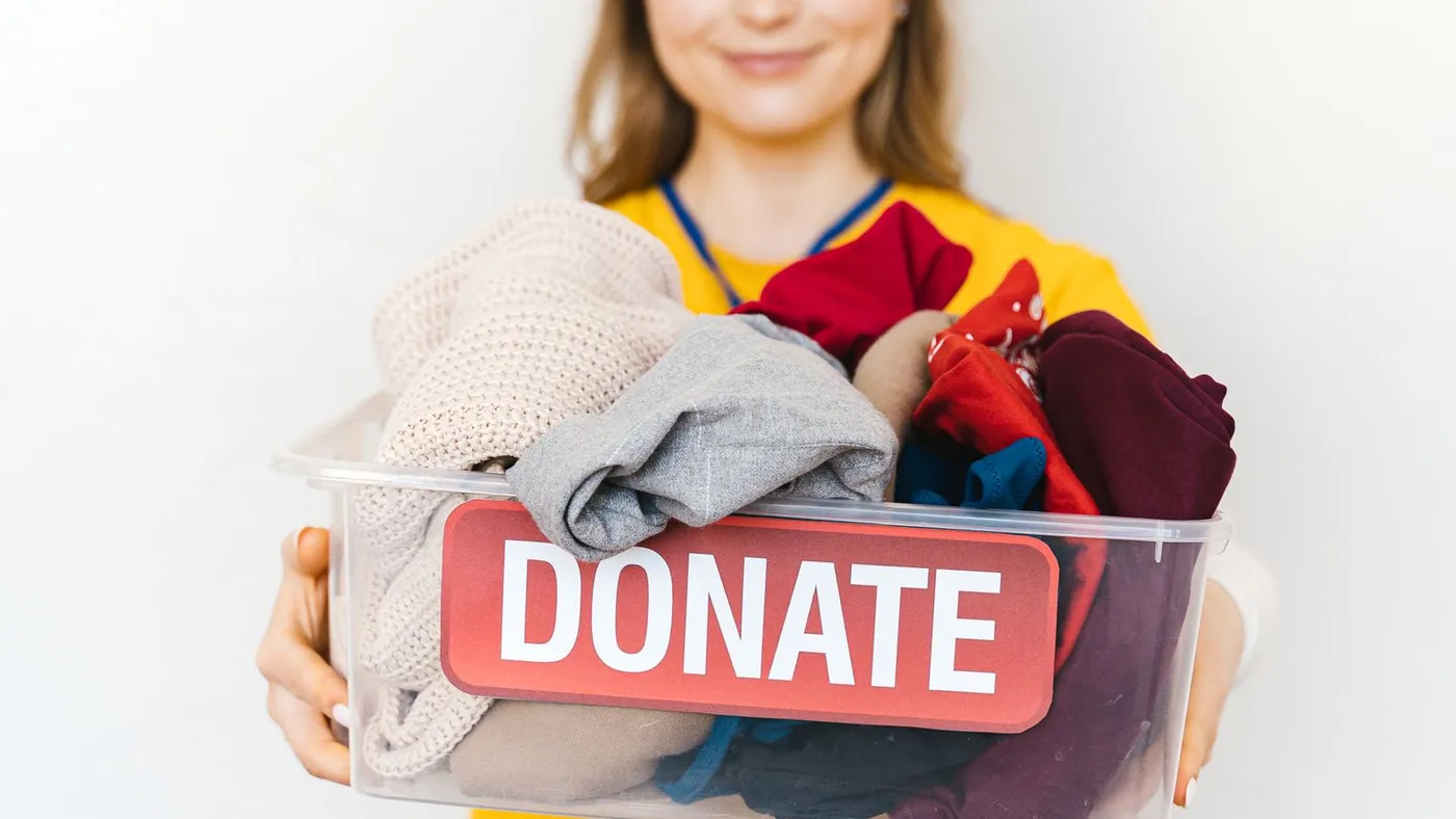 Smiling volunteer in a yellow shirt holding out a clear plastic box labelled Donate and filled with folded clothing
