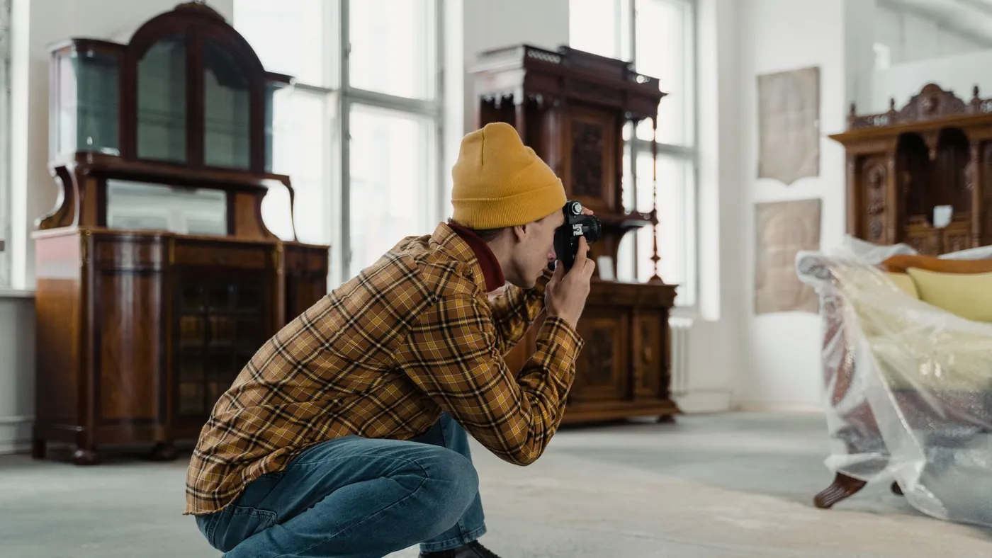 Man in a mustard beanie crouching to photograph a dark wood antique cabinet with a camera, a sofa wrapped in plastic beside him