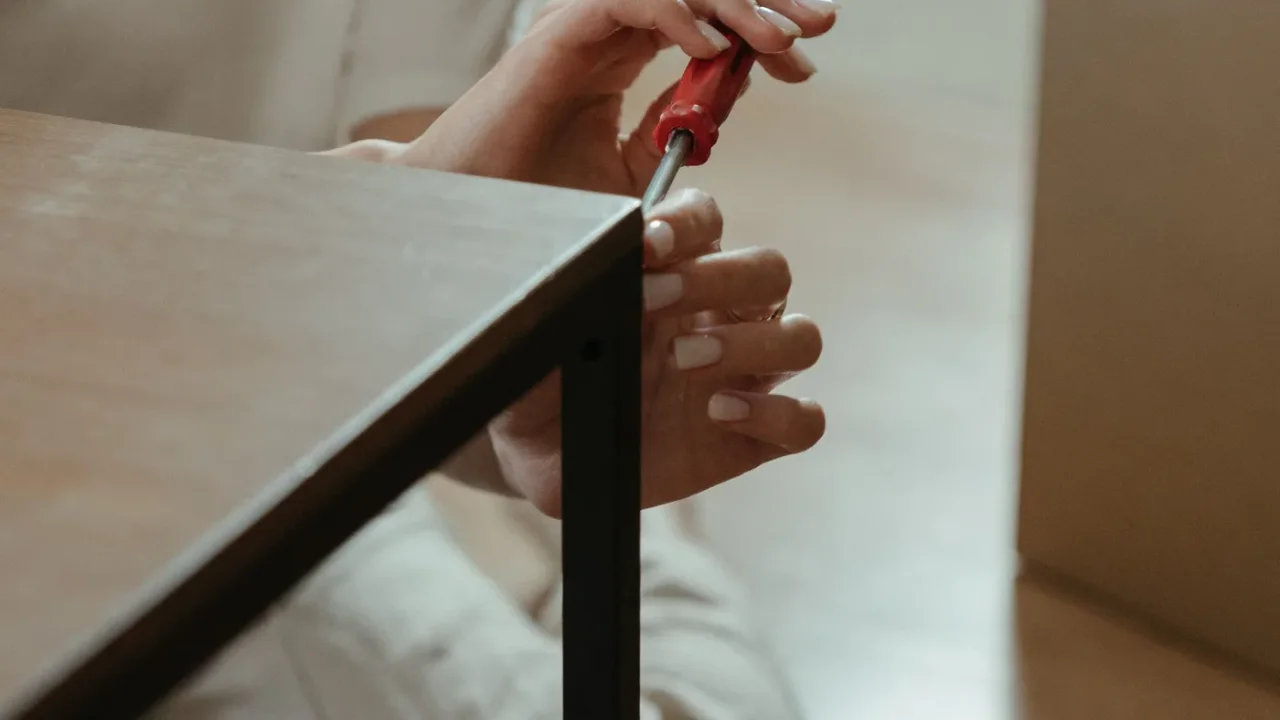 Hands holding a red screwdriver against the black metal frame of a wooden-topped table