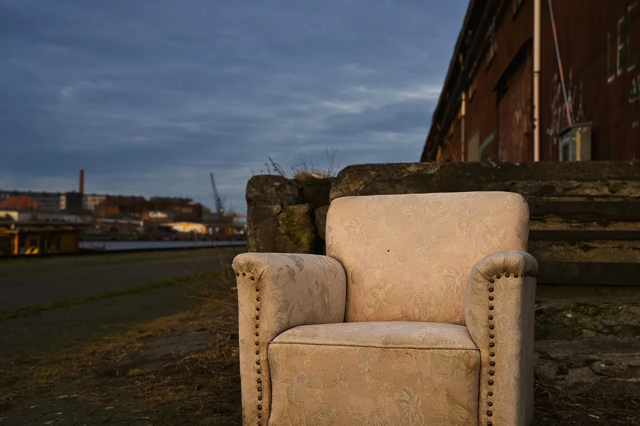 Worn cream floral armchair standing alone on rough ground beside weathered concrete steps with an industrial harbour in the background