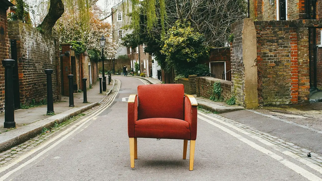 Single red upholstered armchair with pale wooden legs standing alone in the middle of an empty residential street lined with brick walls