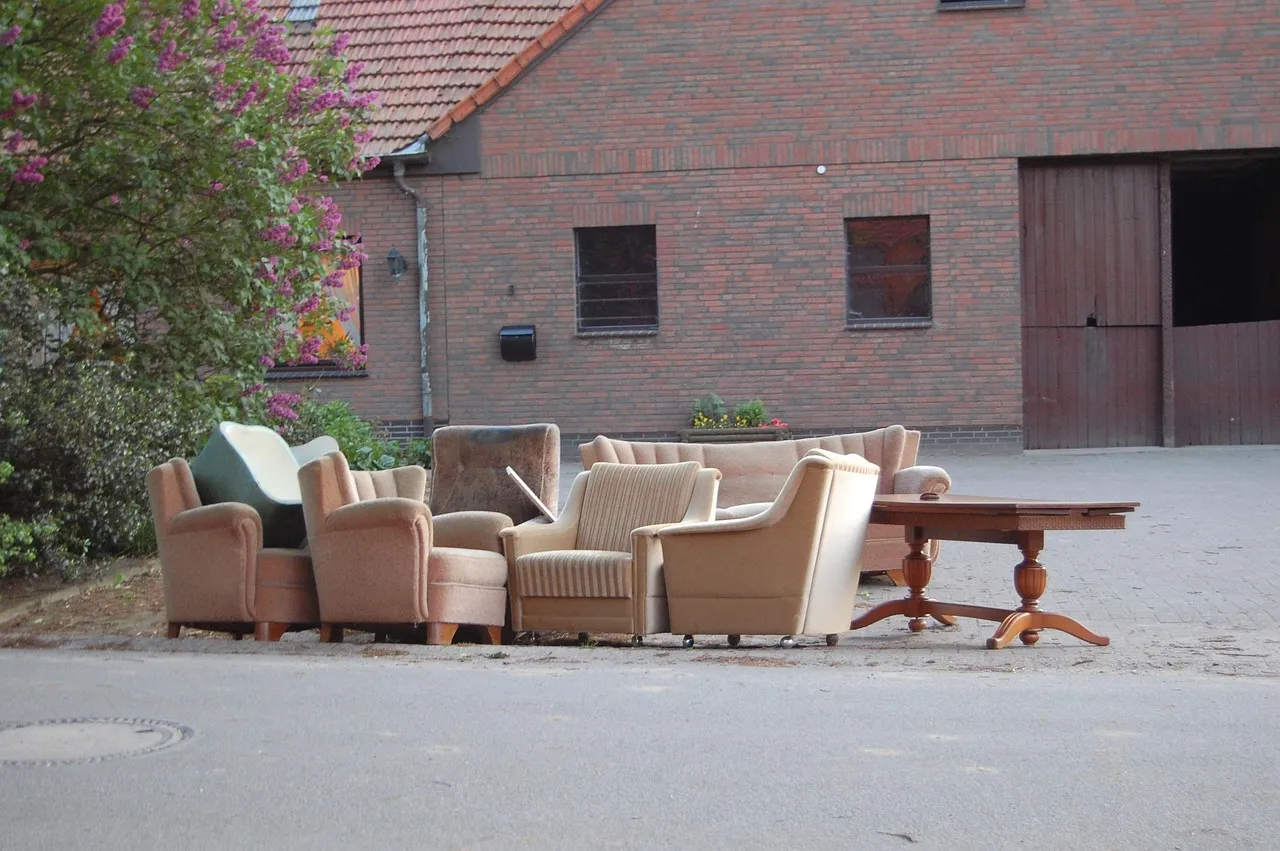 Six worn fabric armchairs and a dark wooden dining table set out together on a paved area in front of a brick building