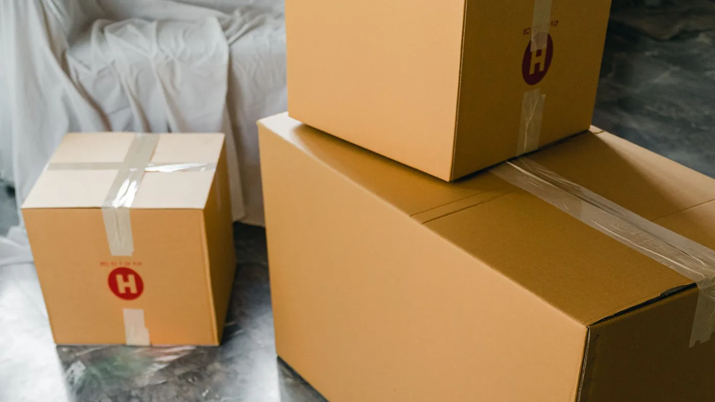 Three sealed brown cardboard moving boxes taped shut and stacked on a floor beside a white dust sheet