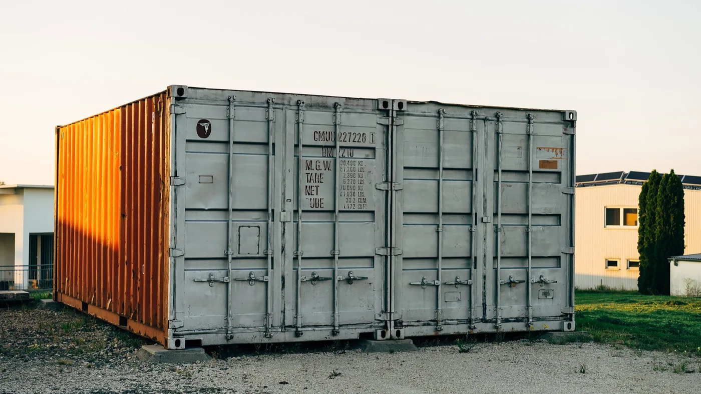 Grey steel shipping container with an orange container behind it standing on grass at an outdoor storage yard