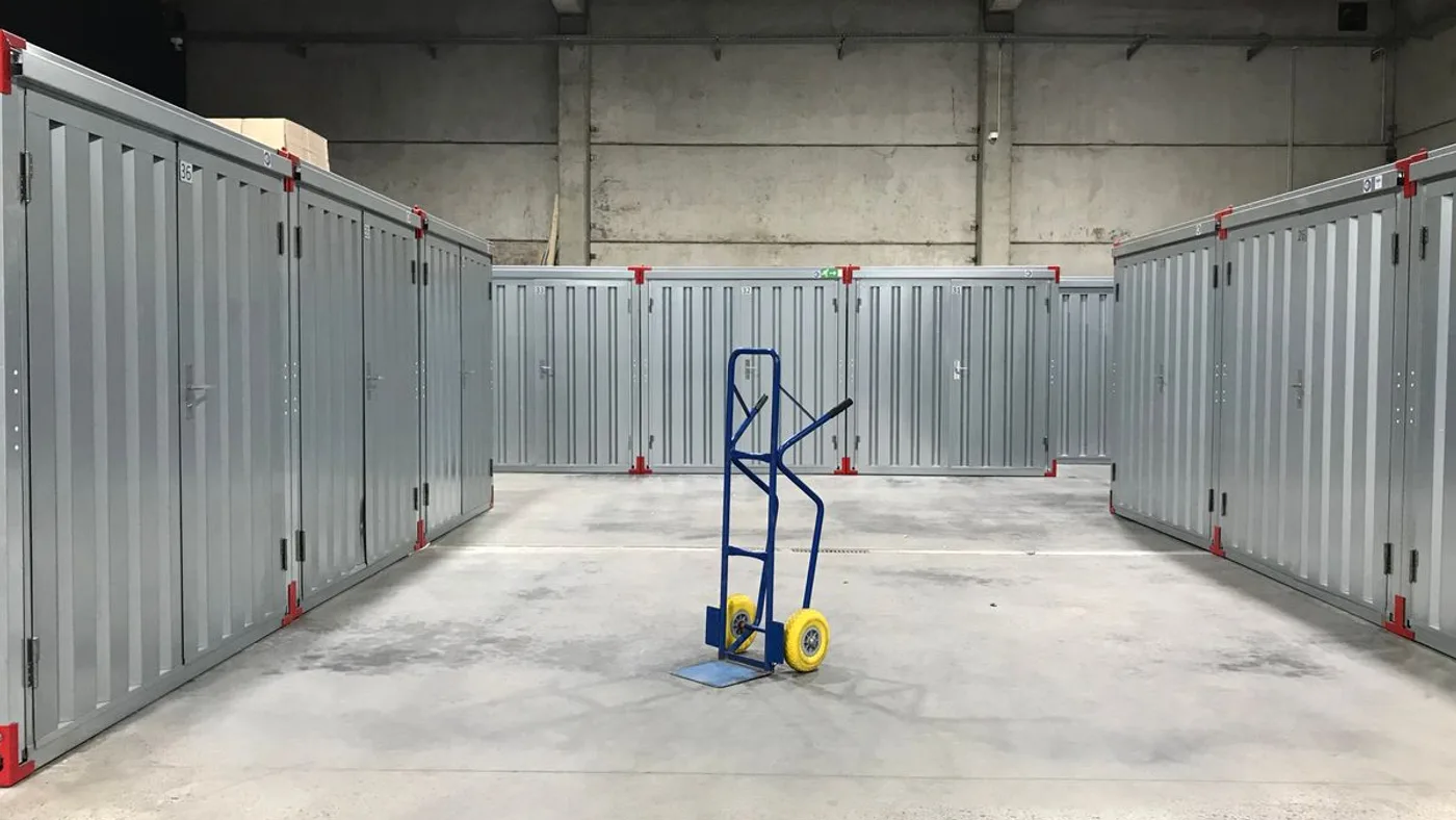 Wide aisle between two rows of grey metal self storage units with a single blue hand trolley parked in the middle of the concrete floor