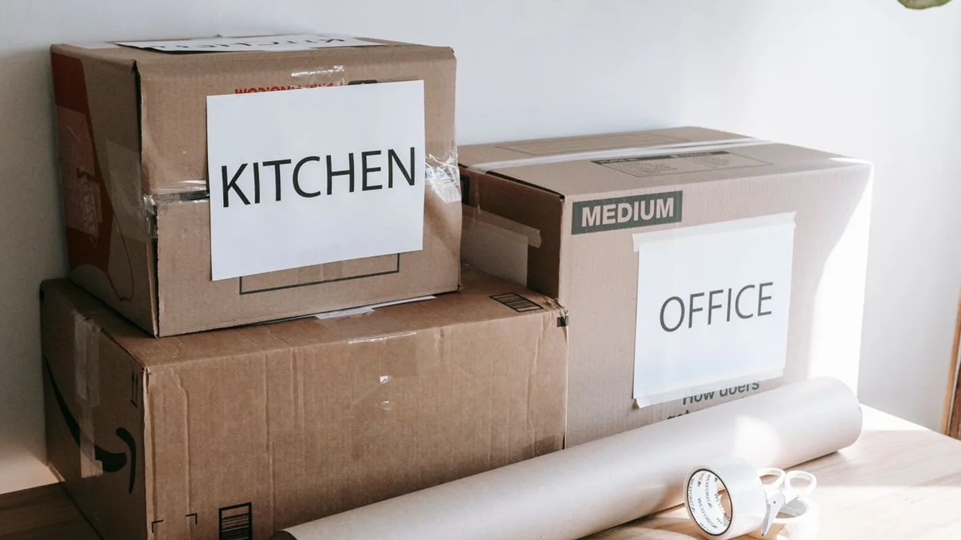 Two cardboard cartons labelled KITCHEN and OFFICE on handwritten signs, stacked beside a roll of packing paper, tape and scissors