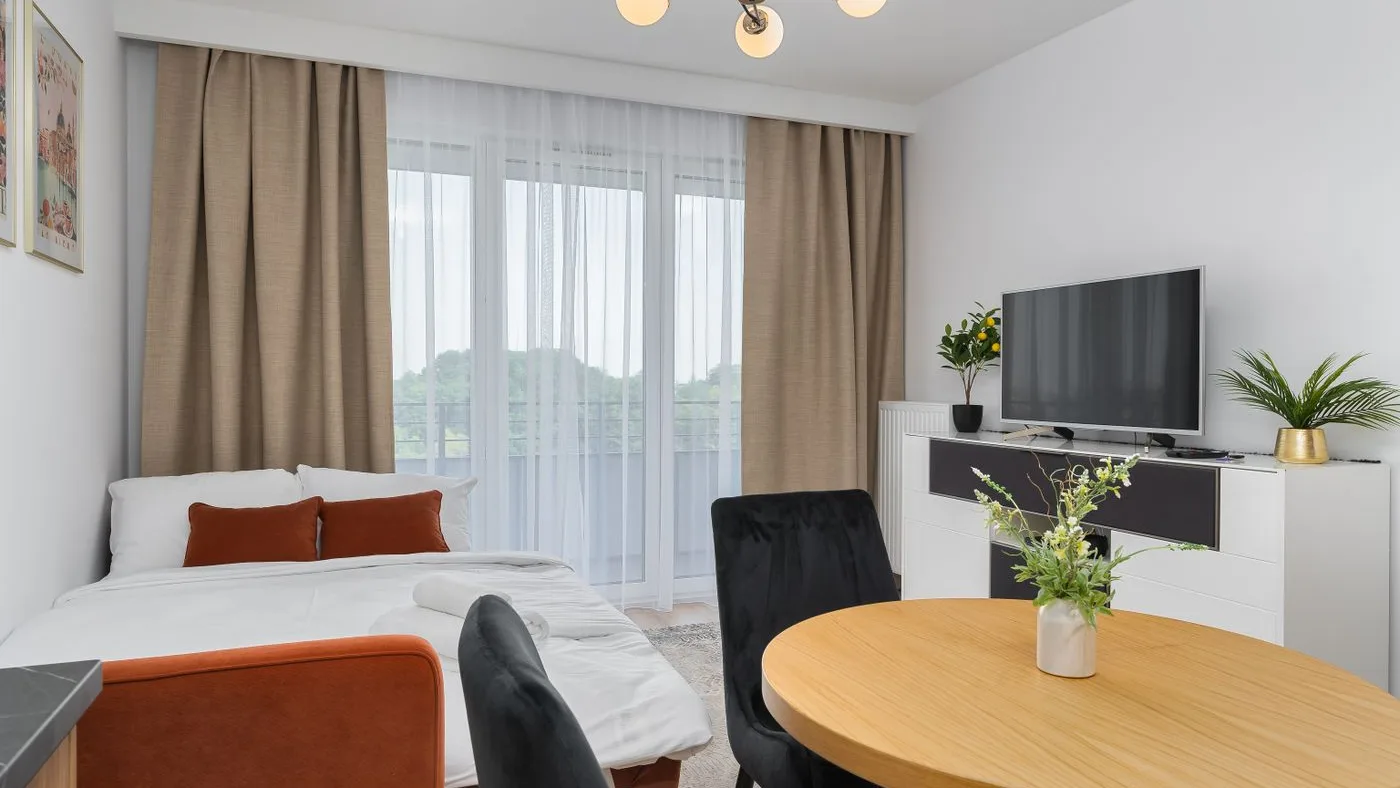 Studio apartment with a double bed, round wooden dining table, dark chairs and a wall-mounted television all in one room