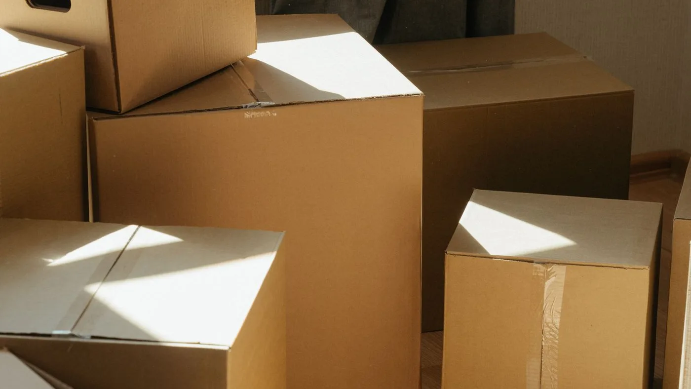 Sunlit floor covered with packed cardboard boxes of several sizes stacked beside a window and a grey curtain