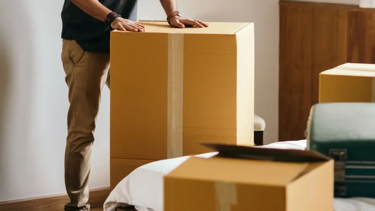 Hands resting on a tall taped cardboard box in a bedroom, with a teal hard-shell suitcase and an open box on the bed beside it
