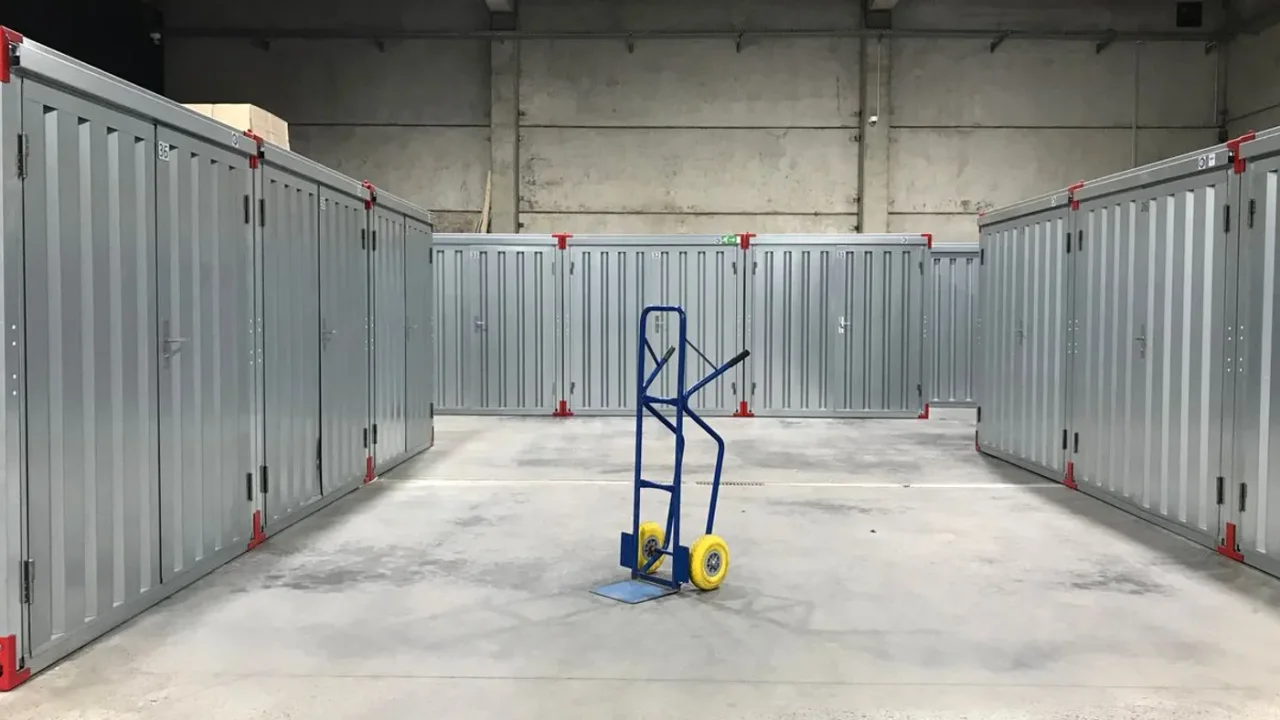 Aisle between rows of grey metal self storage units in a warehouse, with a blue hand trolley standing on the concrete floor