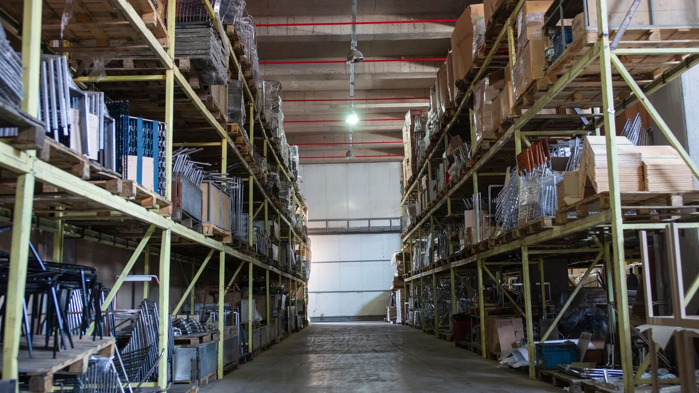 Warehouse storage aisle with metal furniture frames and boxed components stacked on tall industrial racking