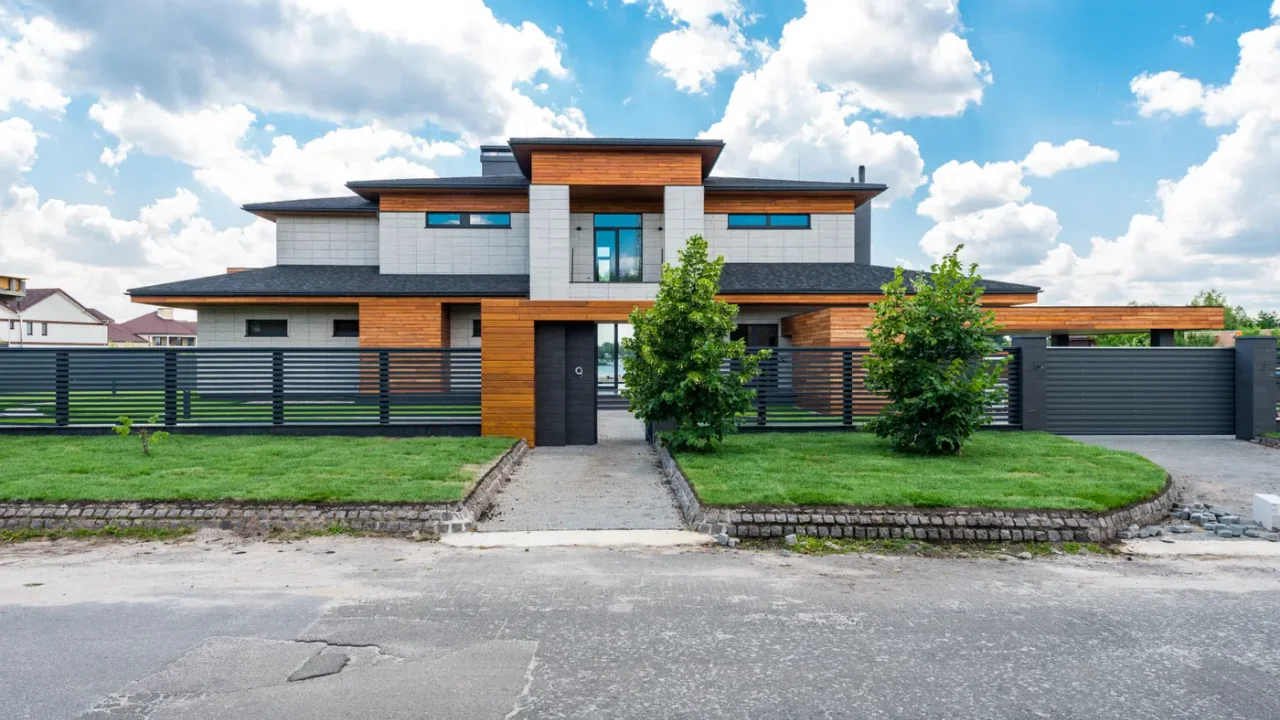 Modern two-storey villa with timber cladding behind a low fence, with a paved path between two small trees and green lawns in front