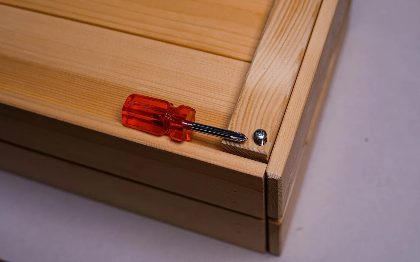 Short red-handled screwdriver resting on the corner joint of a wooden panel unit beside a partly removed screw