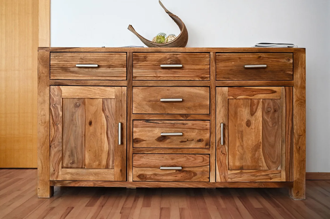 Low solid-wood media sideboard with three drawers, two cupboard doors and bar handles standing against a white wall on a wood floor
